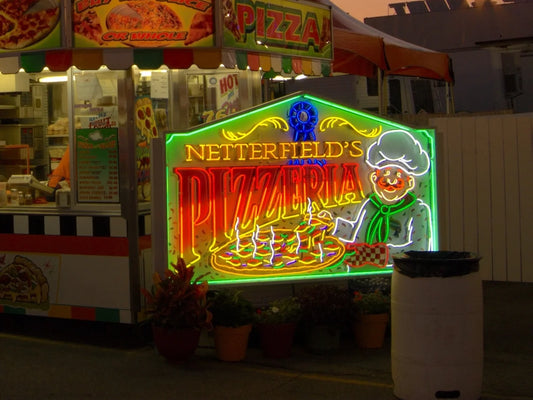 Neon sign for Netterfield's Pizzeria with a smiling chef holding a pizza, in front of a food stall.