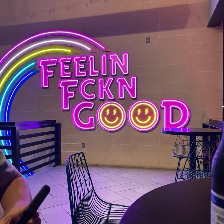 Neon sign reading "FEELIN FCKN GOOD" with two yellow smiley faces and a multicolored rainbow on a tiled wall, with black wireframe chairs and tables.