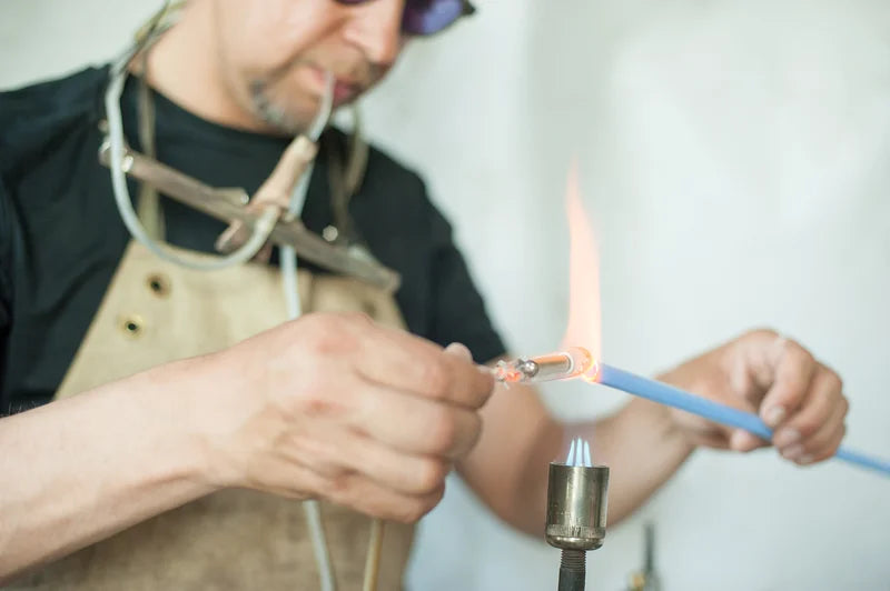 Craftsman using a blowtorch to heat and shape a blue glass tube