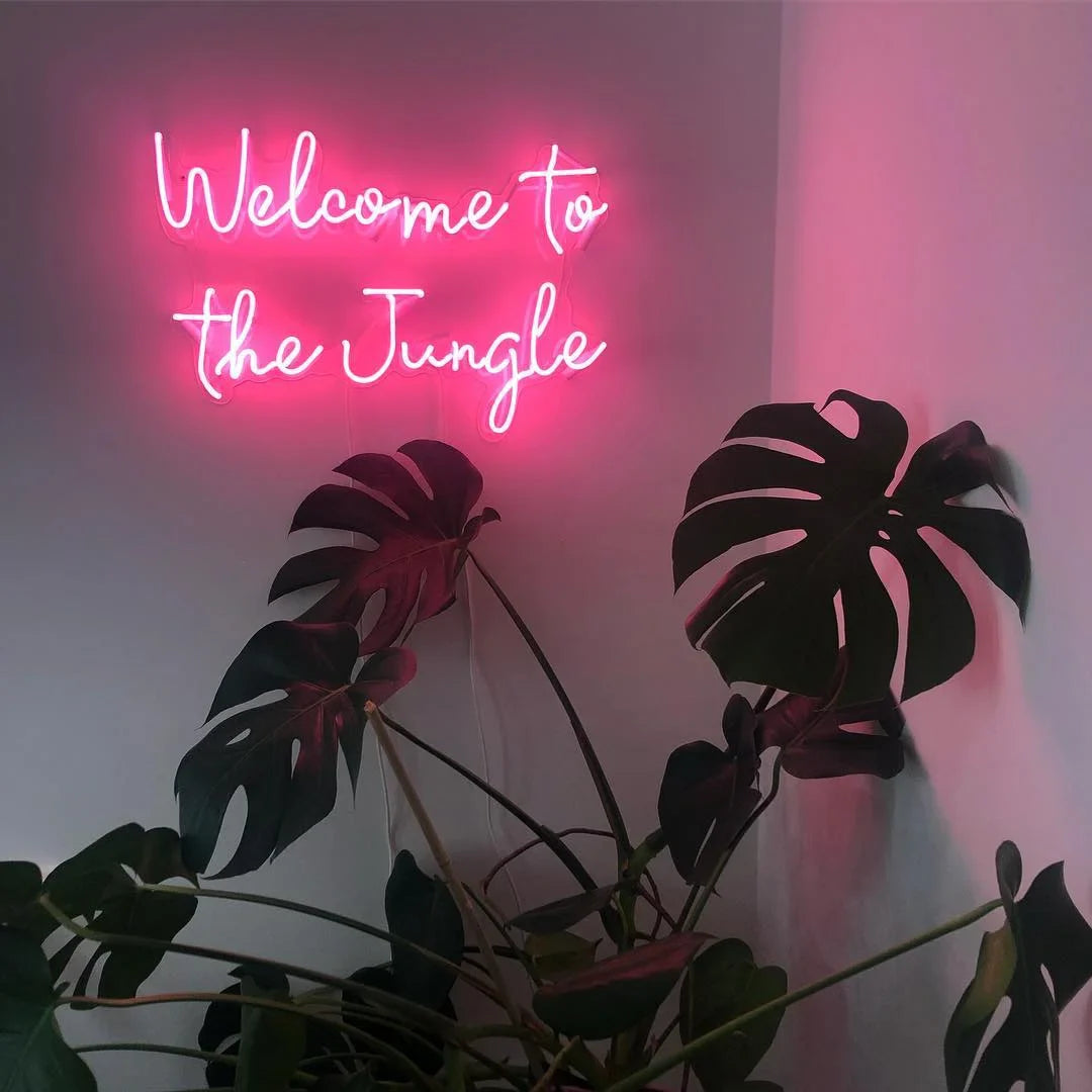 Pink neon sign reading "Welcome to the Jungle" in cursive script with green monstera leaves below it.